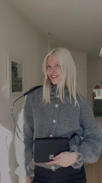 Šedý pletený svetr s velkými knoflíky, doplněný o malou černou koženou kabelku s logem. Doplňky zahrnují tenký pásek.