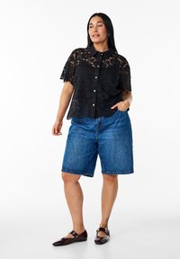Sort blonde bluse med kortærmer og knaplukning, parret med blå denimshorts og mørkkfarvede flade sko. Har blomsterlacet mønster og en afslappet pasform.