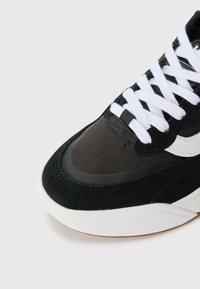 Baskets noires combinant des matériaux en daim et textile, lacets blancs, et une semelle extérieure en caoutchouc blanc texturée avec un accent marron.