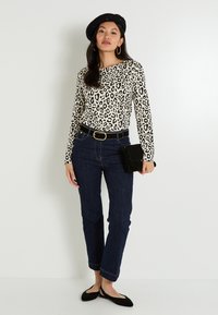 Haut à manches longues en imprimé léopard, jeans bleu marine, ceinture noire avec boucle dorée, béret noir et pochette noire. Des chaussures plates noires complètent la tenue.