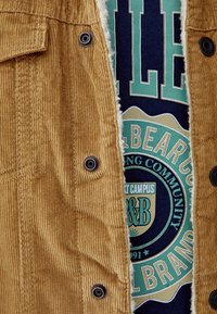 Bruine corduroyjas met een zachte voering, voorzien van zwarte drukknopen. Daaronder toont een marineblauw t-shirt een kleurrijke grafische print en tekst.