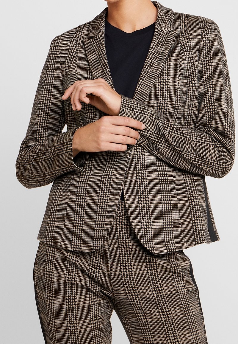 Blazer à carreaux marron et noir avec une coupe ajustée, doté de revers crantés et de manches longues, associé à un top noir uni en dessous.
