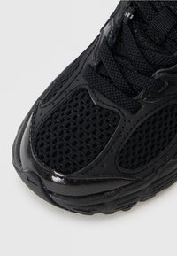 Chaussure de sport noire avec une tige en mesh, des accents brillants lisses, une semelle épaisse et une semelle en caoutchouc texturée avec des coutures détaillées et un laçage.