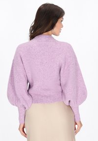 Pulover tricotat texturat în nuanță de liliachiu, cu guler înalt și mâneci tip balon. Dispune de manșete și tiv cu elastic. Stilizat cu o fustă bej.