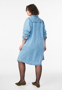 Svetlomodrá denimová šaty košeľa s vyšívanými kvetinovými akcentmi, dlhými rukávmi a zakriveným lemom. Nosené s tmavými pančuchami a hnedými balerínami.