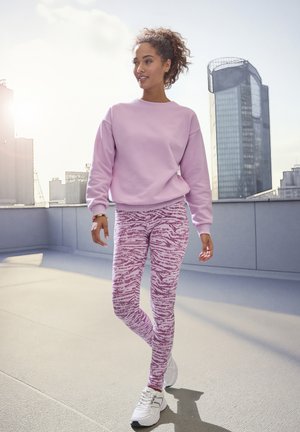 Hellrosa Sweatshirt mit gerippten Bündchen, kombiniert mit pink-weißen Leggings im Tigerstreifen-Muster, abgerundet mit weißen Sneakers.