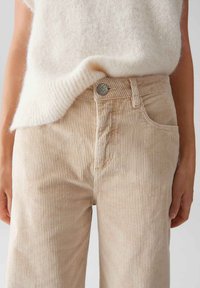 Beige Corduroyhosen mit vertikalen Rippen, ausgestattet mit einem silbernen Knopf an der Taille, Fronttaschen und einer weichen Textur. Kombiniert mit einem leichten Pullover.