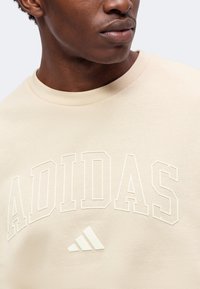 Κοντινό σε άτομο που φοράει μπεζ φούτερ Adidas με ανάγλυφο όνομα εμπορικού σήματος και λογότυπο στο στήθος.