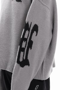 Grijze gebreide trui met een gestructureerde afwerking en zwarte grafische accenten op de mouw. Geribbelde boorden en zoom zorgen voor structuur.