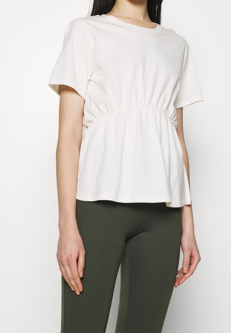 Femme portant un haut blanc ample à manches courtes avec un détail froncé à la taille et un pantalon ajusté vert olive, debout devant un fond blanc uni.