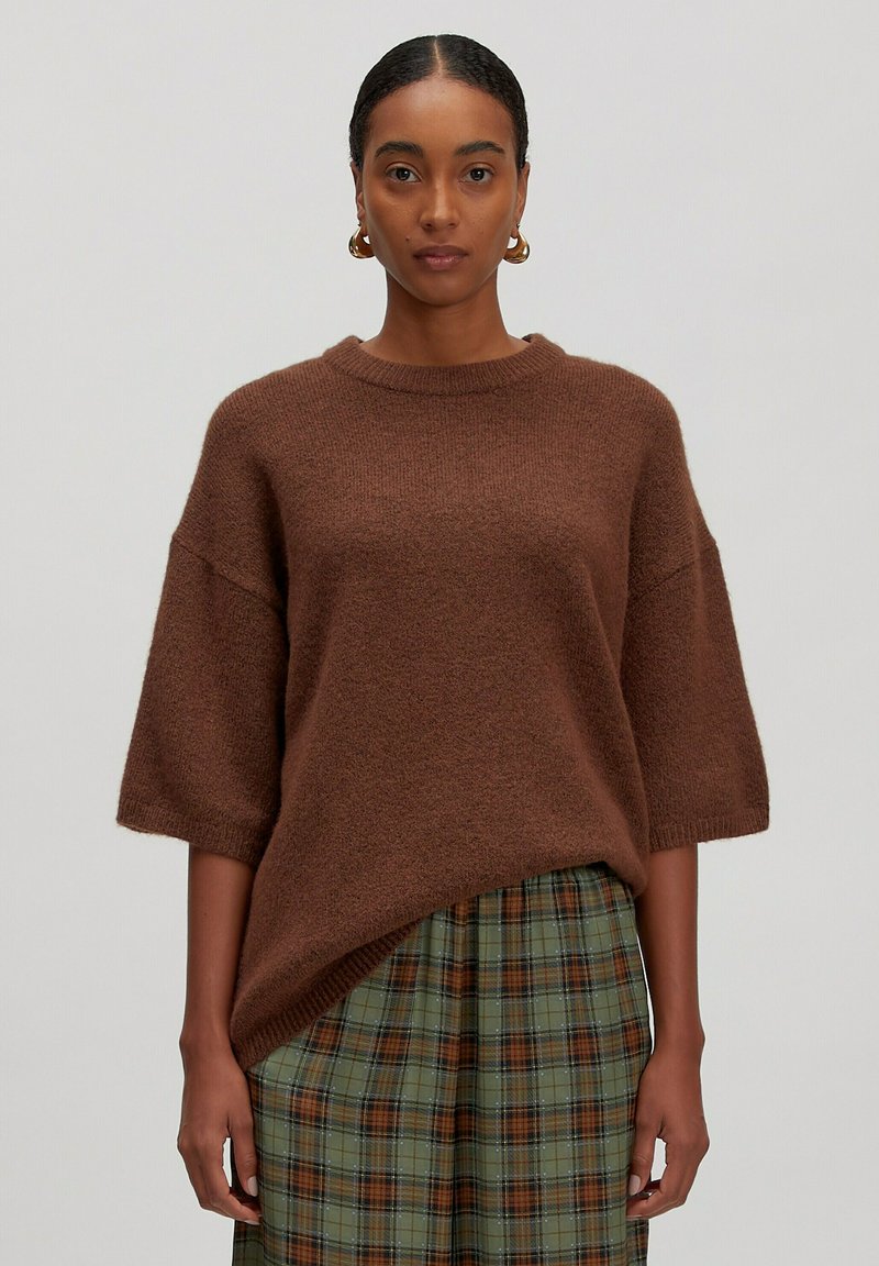 Brauner übergroßer Pullover mit kurzen Ärmeln, aus strukturiertem Strickmaterial, kombiniert mit einem grün karierten Rock mit braunen und blauen Akzenten.