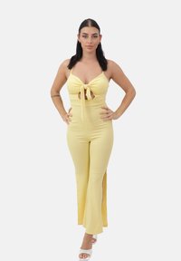Mono amarillo de canalé con cuello halter y detalle de lazo en el frente. Piernas acampanadas con aberturas laterales, combinado con sandalias de tacón blanco con tiras.