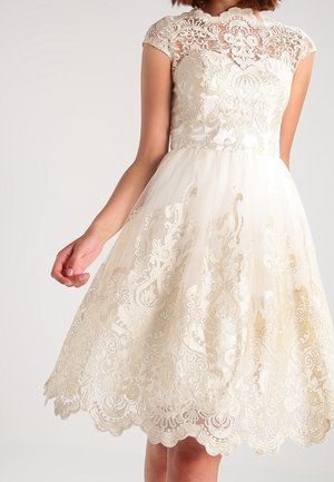 Kvinde iført hvid blondekjole med kappelignende ærmer og blomstret broderi, tætsiddende krop og udvidet nederdel med bølget kant.