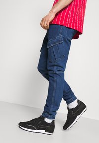 Pantalones cargo de mezclilla en azul oscuro con puños elásticos en los tobillos y bolsillos laterales. Combinados con zapatillas negras y calcetines blancos. Camisa de patrón rojo visible.