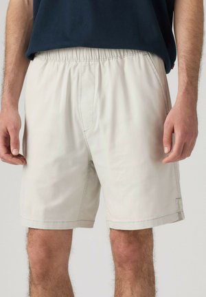 Mann trägt hellbeige elastische Taillenshorts und ein dunkelblaues Hemd, Vorderansicht von der Taille bis zu den Knien.