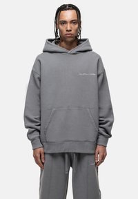 Felpa grigia in morbido tessuto con tasca a marsupio, cappuccio con cordino e cuciture a contrasto; abbinata a pantaloni della tuta coordinati.