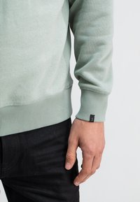 Lichtgroene sweatshirt met ribgebreide manchetten en zoom, voorzien van een klein zwart label op de mouw. Gekozen in combinatie met donkere, gestructureerde broek.