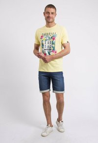 T-shirt jaune à manches courtes avec des motifs floraux et le texte "PARADISE", associé à un short en denim retroussé et des baskets blanches.
