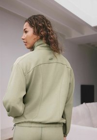 Světlezelená mikina s vysokým límcem, žebrovaným lemem a jemným prošíváním. Na zadní straně blízko výstřihu se nachází logo "self.".