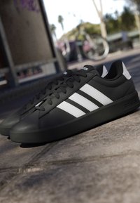 Baskets noires Adidas avec des bandes blanches et un logo sur le côté, placées sur une surface en béton à l'extérieur avec un vélo flou en arrière-plan.