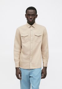 Beige skjorte med knapper foran, to lommer foran, metallknapper og lange ærmer, serveret med lyseblå jeans.
