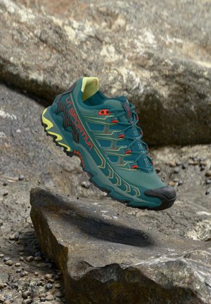 Scarpe da corsa turchesi con dettagli neri e gialli, tomaia in rete, suola testurizzata e design aerodinamico. Presenta un sistema di allacciatura prominente e un logo.