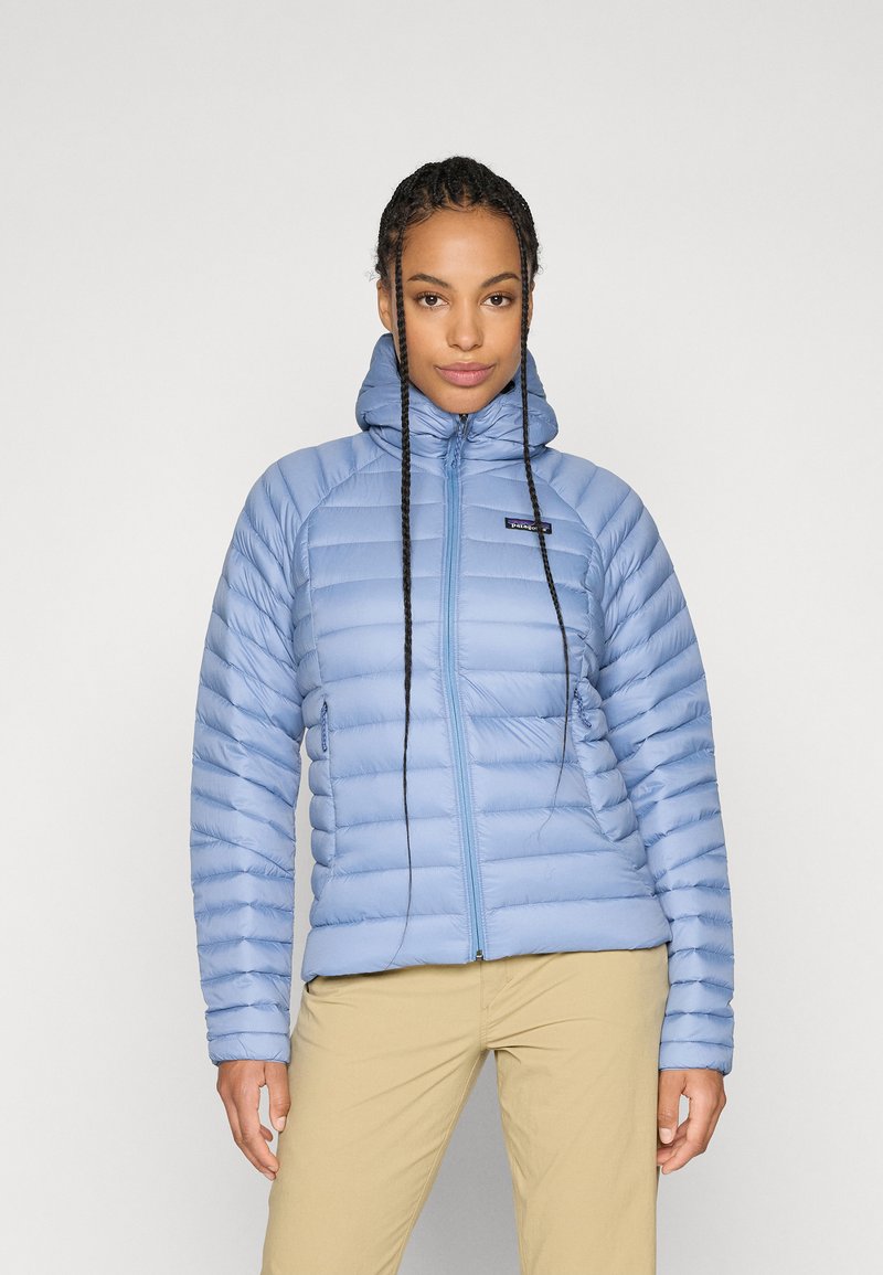 Žena s pletenom kosom koja nosi svijetloplavu puffer jaknu i bež hlače, stoji pred jednostavnom svijetlom pozadinom.