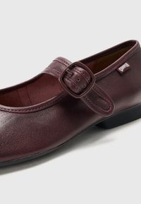 Burgundy leather shoe with a rounded toe, adjustable strap with a buckle, decorative stitching, and a low black heel.