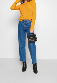 Felgele geribbelde trui gecombineerd met een hoge taille blauwe spijkerbroek. Model draagt een kleine zwarte handtas met een textuur. Zwarte enkellaarsjes met een vierkante neus.