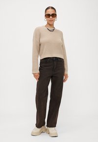Beige gebreide trui met een loose fit, brede manchetten, donkerbruine rechte jeans en stevige beige sneakers. Zwarte zonnebril en een gelaagde ketting.
