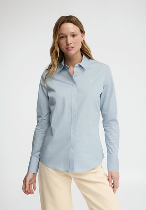 Vrouw met lichtbruin haar, gekleed in een lichtblauw overhemd met knopen en beige broek, tegen een effen witte achtergrond.