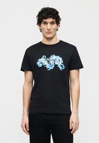 Νέος άνδρας με σγουρά μαλλιά που φοράει μαύρο μπλουζάκι με μπλε κυματιστό σχέδιο και λευκή επιγραφή "Versace Jeans Couture" στο μπροστινό μέρος.