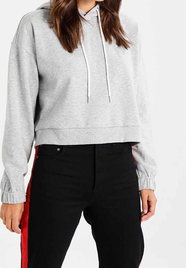 Donna che indossa un hoodie corto grigio chiaro con lacci bianchi e pantaloni neri con strisce laterali rosse, in piedi contro uno sfondo neutro.