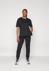 Svart kortermet topp med et teksturert geometrisk mønster, kombinert med svarte joggebukser. Modellen har på seg hvite joggesko med svarte detaljer.