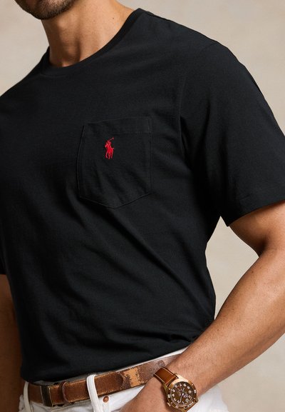 Moški, ki nosi črno polo majico z logotipom in prsno žeprico, rjav usnjen pas, bele hlače in rjavo ročno uro na levi roki.