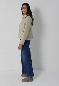 Beige, geprofileerde jas met een kraag en twee voorzakken, gecombineerd met ruime blauwe jeans en grijze schoenen. Simpel, neutraal kleurenpalet.