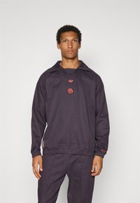 Σκούρα μοβ φούτερ με μεγάλο γιακά, ριμπ μανσέτες και ζώνη στη μέση. Διαθέτει κόκκινα κεντημένα λογότυπα στο στήθος και στον επάνω γιακά.