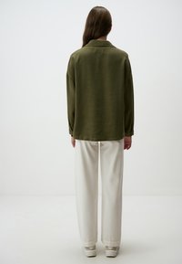 Olijfgroene oversized shirt met een kraag, lange mouwen en een relaxte pasvorm, gecombineerd met witte wijde broek en lichte schoenen.