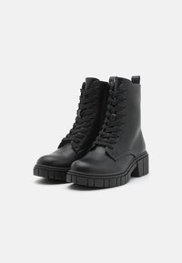 Botas de cuero negro hasta el tobillo con una superficie texturizada, cordones en la parte delantera, cremallera lateral y suela de goma gruesa. El diseño presenta una punta redondeada.