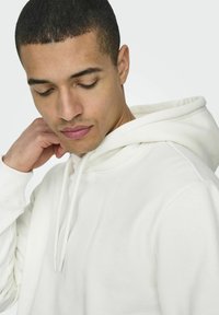 Weißer Kapuzenpullover aus weichem Stoff, mit einer Kängurutasche und Kordeln am Kragen. Glatte Textur, lockere Passform.
