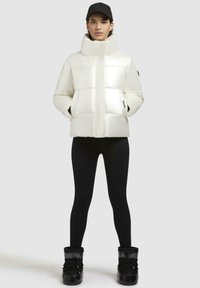 Giacca bianca piumosa con colletto alto e finitura lucida, dotata di due tasche laterali, abbinata a leggings neri e stivali invernali scuri.