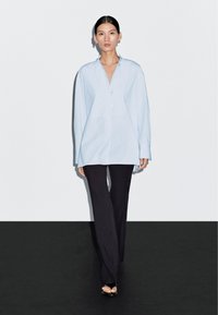 Light blue oversized shirt with a collar, long sleeves, and a button front, paired with black fitted pants and open-toe black sandals.