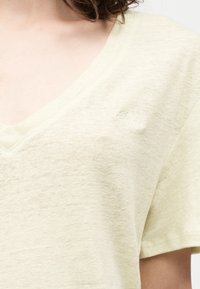 Κοντινό πλάνο ενός ατόμου που φοράει φωτεινό μπεζ λινό ή βαμβακερό μπλουζάκι με V-λαιμόκοψη, κοντά μανίκια και διακριτικό κεντημένο κείμενο στο στήθος.