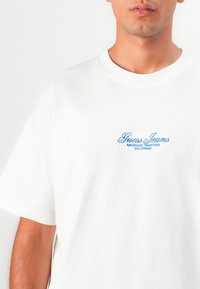 Weißes Baumwoll-T-Shirt mit blauer bestickter Aufschrift "Guess Jeans American Tradition California", mit Rundhalsausschnitt und kurzen Ärmeln.