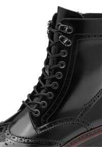 Botte brogue en cuir noir poli avec lacets noirs, œillets métalliques et perforations décoratives sur l'empeigne et le bord de la semelle.