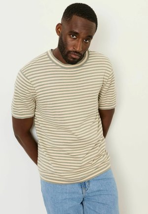 Mann mit Bart, der ein beige-weiß gestreiftes Kurzarmhemd und hellblaue Jeans trägt, steht vor einer schlichten weißen Wand.