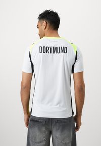 Maglia sportiva bianca con accenti neri e giallo neon, con la scritta "DORTMUND" stampata in grassetto nero sul retro. Tessuto morbido, maniche corte.