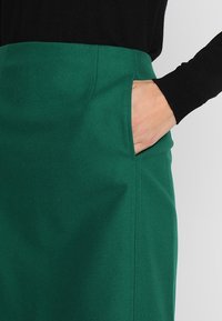 Grøn nederdel lavet af tekstureret stof, med sidelomme og ren syning. Båret med en sort langærmet top.