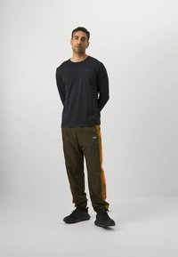 Muž stojaci so založenými rukami, nosí čiernu dlhú košeľu a olivovo zelené nohavice s oranžovými bočnými pruhmi a čierne tenisky.