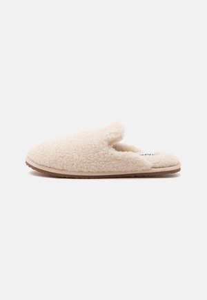 Chaussons beige doux à enfiler avec une surface duveteuse et texturée ainsi qu’une semelle plate marron, conçus pour le confort intérieur.